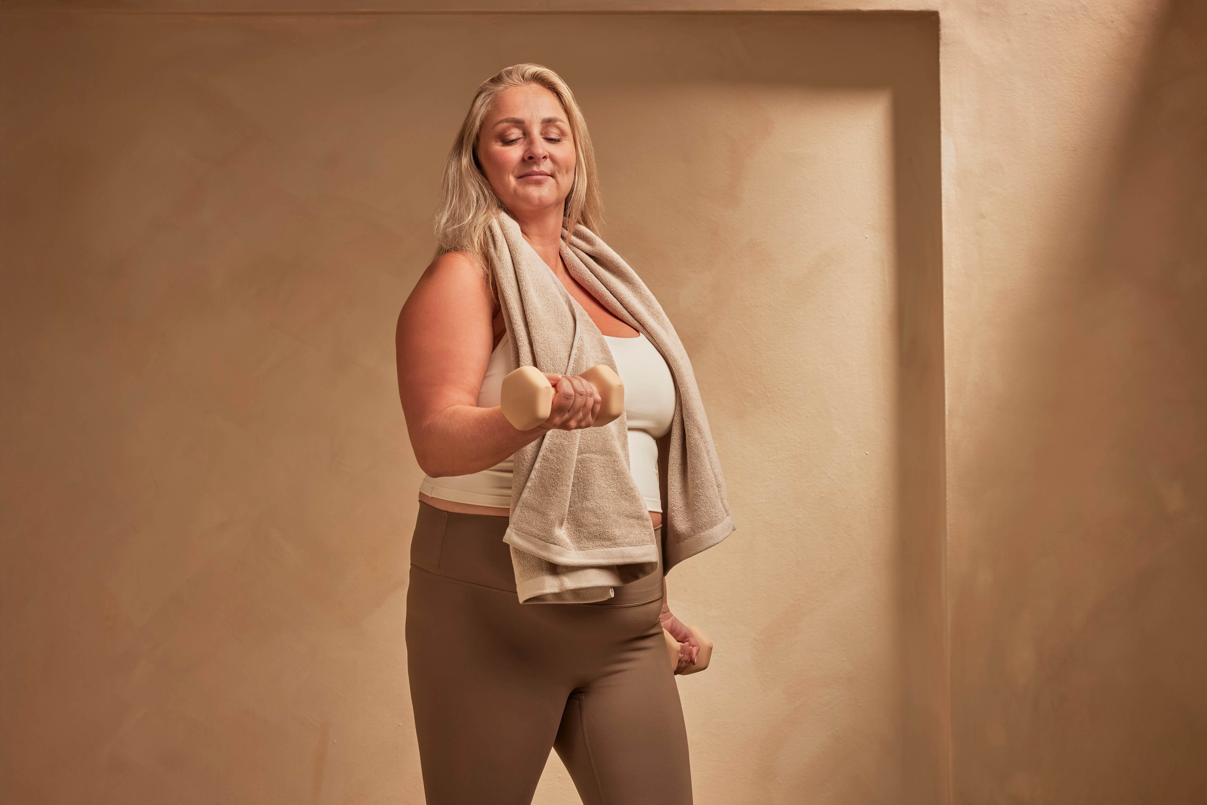 Smiling blonde woman in workout gear lifts a dumbbell with a towel on her shoulder.