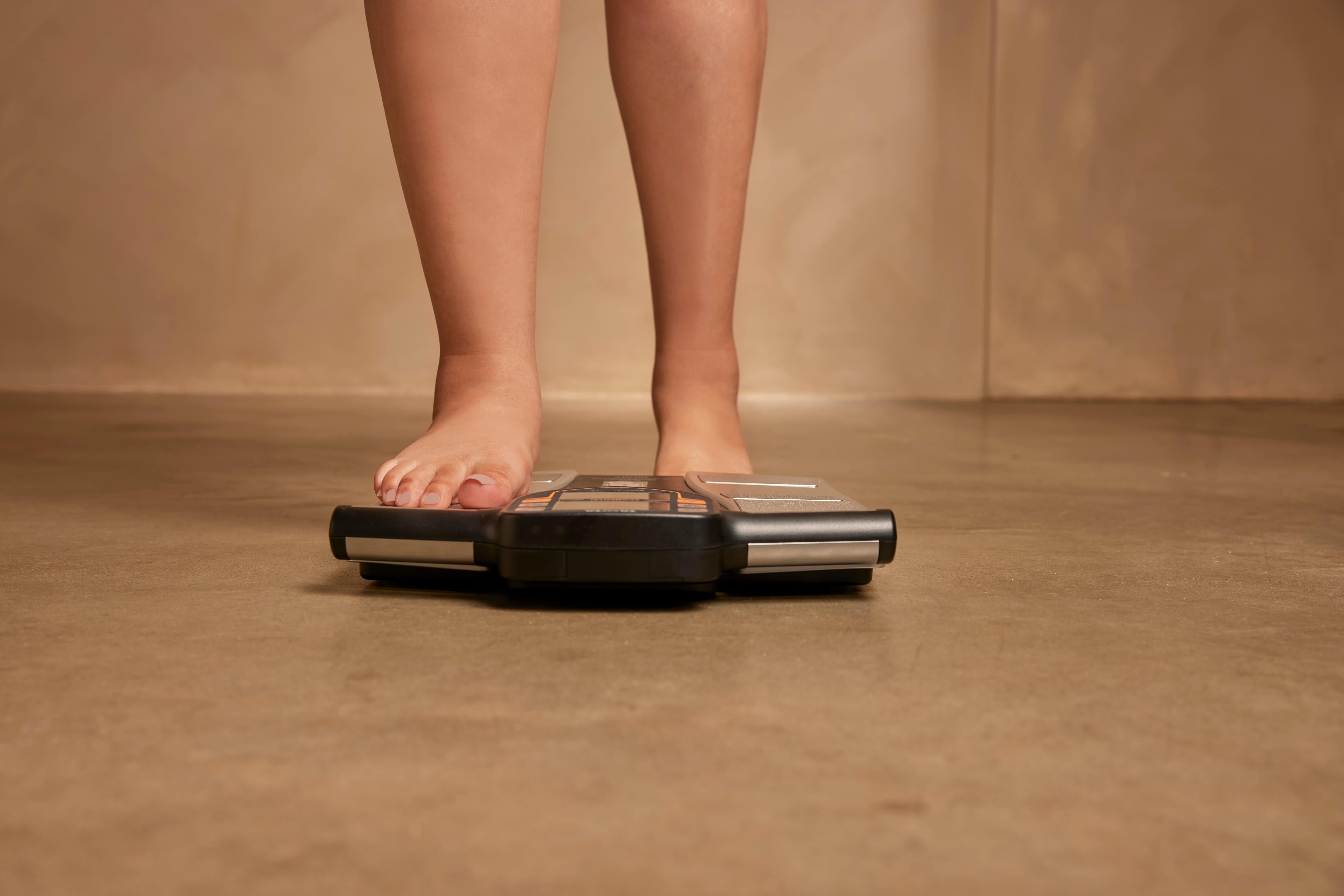 Bare feet stand on a digital weighing scale on a brown floor.