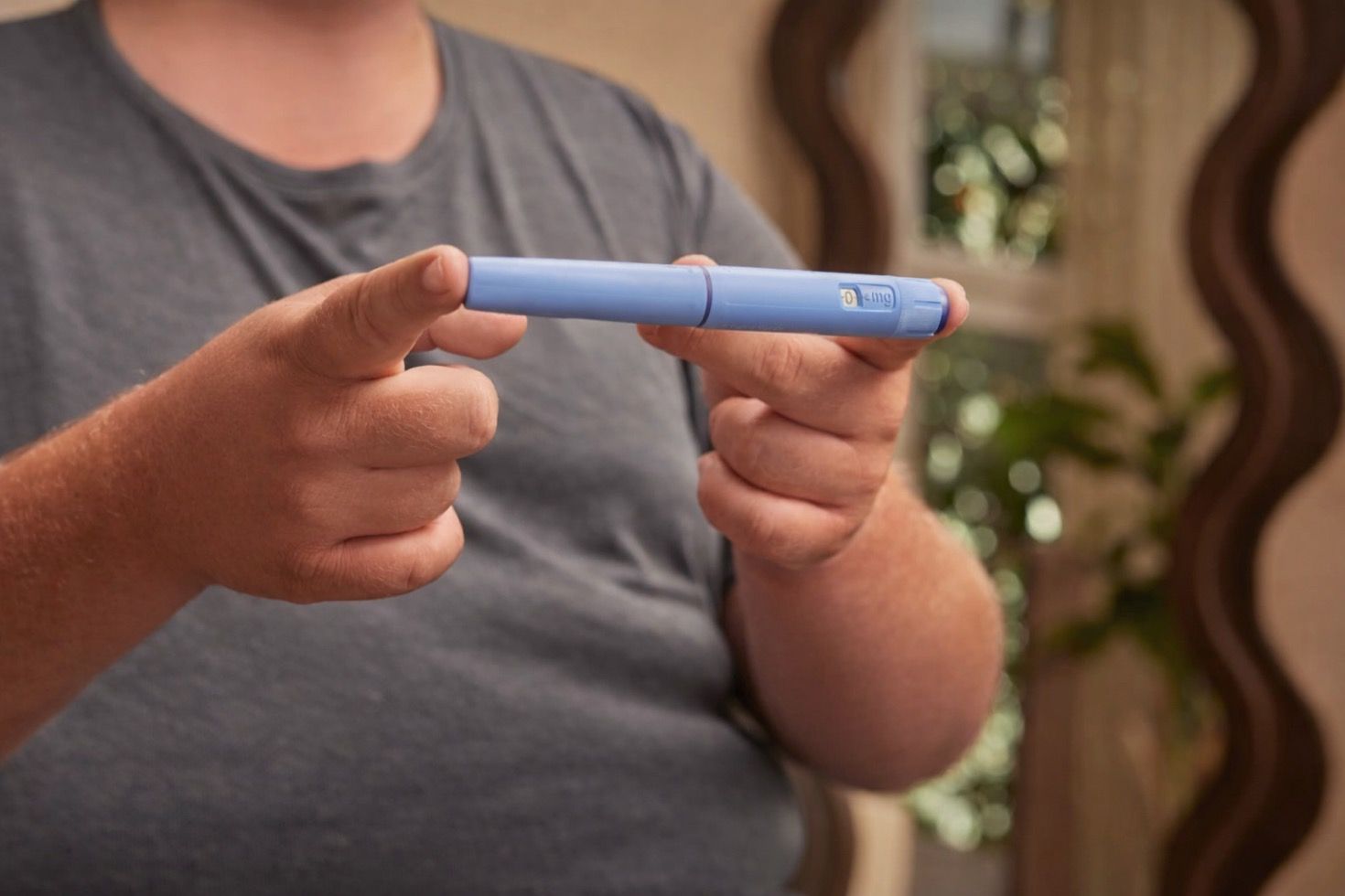 Hands hold a light blue, pen-like medical auto-injector device.